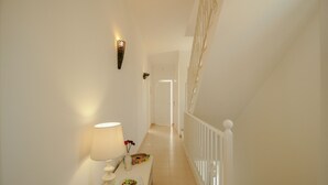 A hallway with a white floor, a white railing, and a white wall. There is a lamp on a table with a vase and a plate.