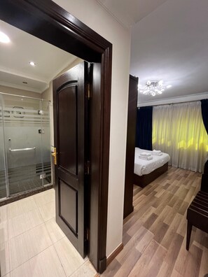 Une chambre d’hôtel avec une porte en bois, un espace de douche en verre, un lit recouvert d’une courtepointe blanche et un lustre.