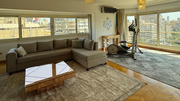 A spacious living room with a large sectional sofa, a coffee table, and a treadmill. The room has large windows offering a city view. - VESTA - Mohandessin (Giza)