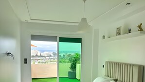 Een moderne slaapkamer met een groot bed, uitzicht op een groen gazon en gebouwen, en een plafondlamp.
