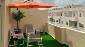 Een balkon met kunstgras, twee lounge stoelen, een oranje parasol en een potplant.
