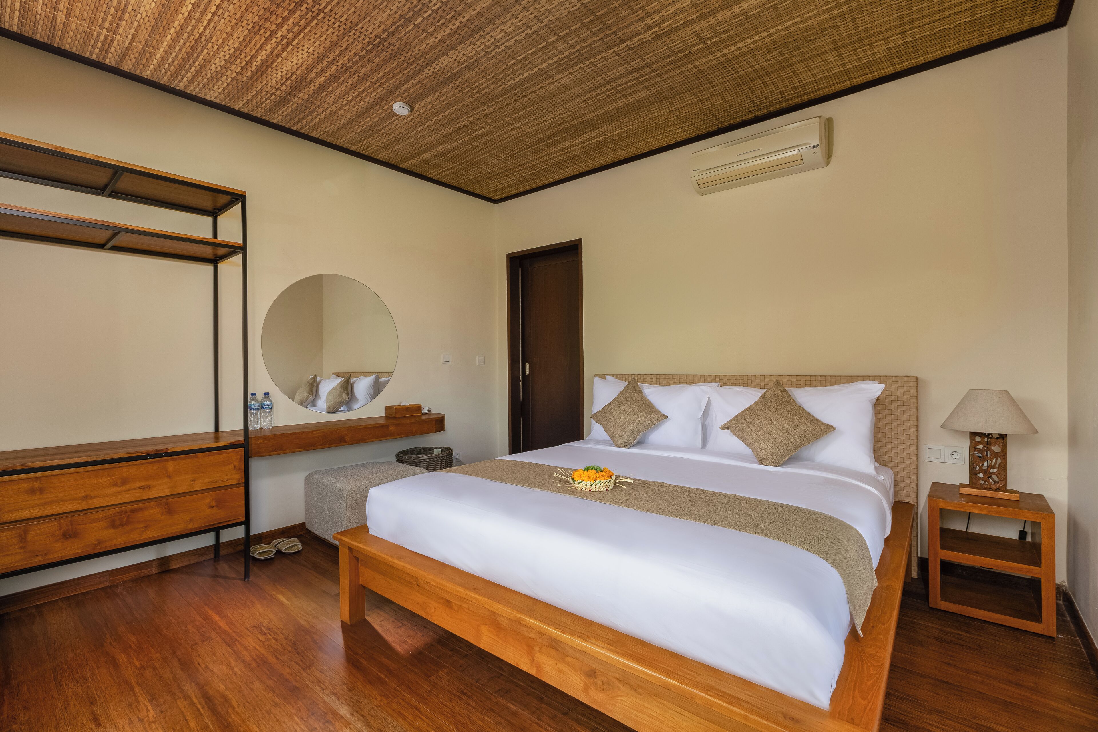A hotel room with a wooden bed, a round mirror, a wooden bench, a small table with a lamp, and a ceiling fan.