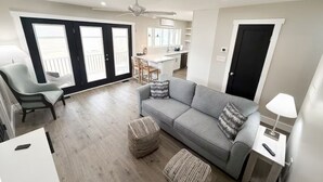A modern living room with a grey sofa, two ottomans, a white side table, and a chair. There is a kitchen area in the background with a dining table and chairs. - 2 Bedroom Riverfront House on the New River Trail (Allisonia)