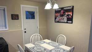A dining area with a white table, four chairs, and patterned placemats. There is a door with a window, a framed picture on the wall, and a ceiling light fixture. - Walk to Downtown CDA | Sleeps 8 | Arcade + Pets (Coeur d'Alene)