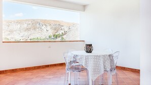 Un'area pranzo con tavolo e sedie, vista su un paesaggio montuoso attraverso una finestra e un pavimento piastrellato.