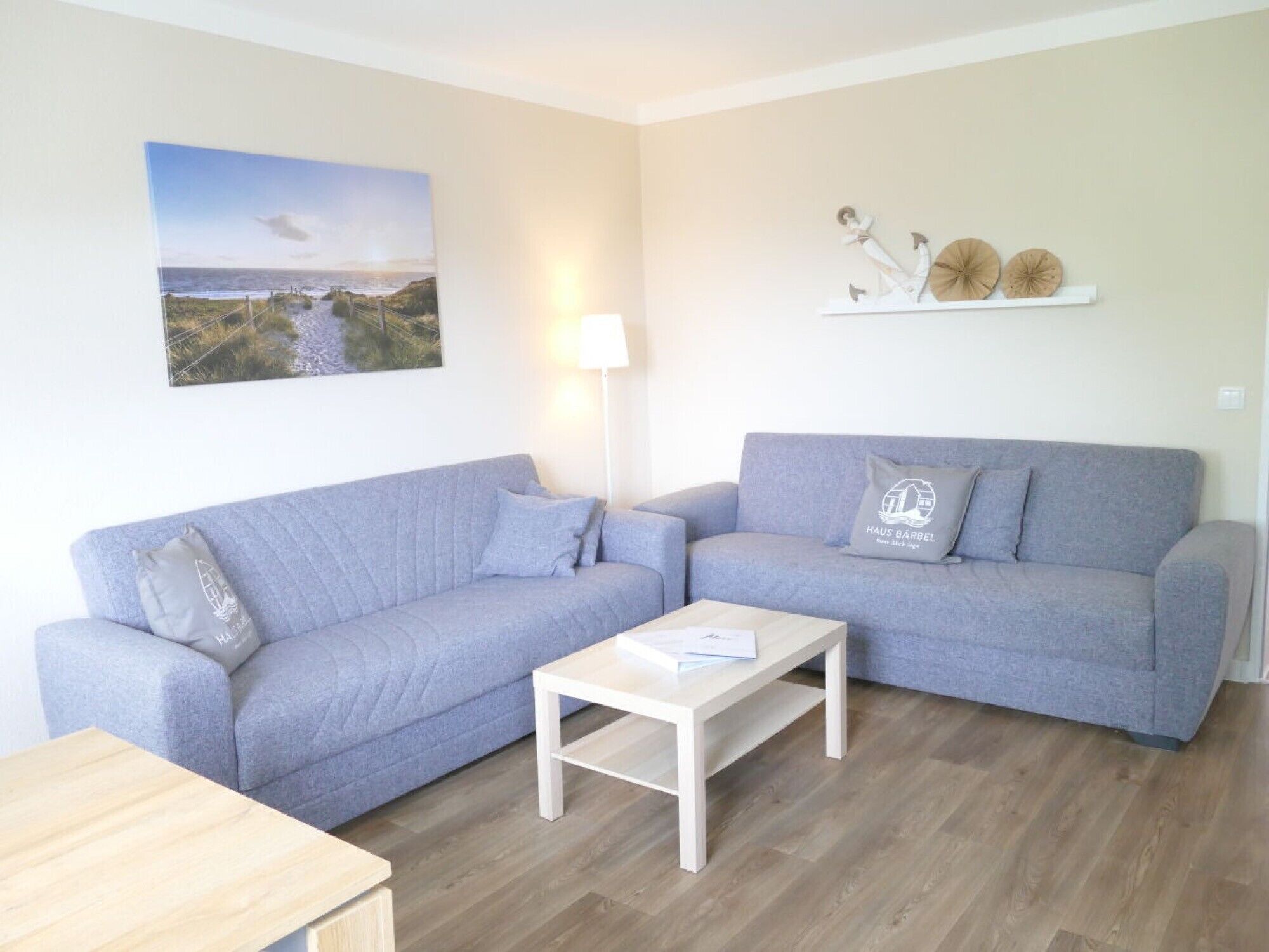 A living room with two grey sofas, a white coffee table, a lamp, a painting of a coastal scene, and decorative items on a shelf.