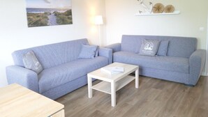 Een woonkamer met twee grijze banken, een witte salontafel, een lamp, een schilderij van een kustlandschap en decoratieve objecten op een plank.