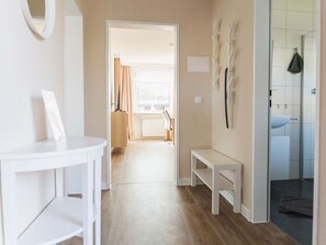 Un couloir avec un banc blanc, un porte-manteau et une salle de bain équipée d’un lavabo et d’une douche.