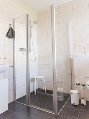 Un baño moderno con ducha de vidrio, inodoro blanco, un taburete pequeño blanco y un cubo de basura.