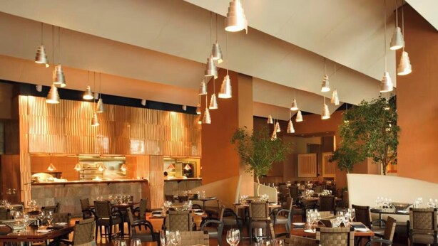 A restaurant with multiple dining tables, hanging pendant lights, and a kitchen area in the background.