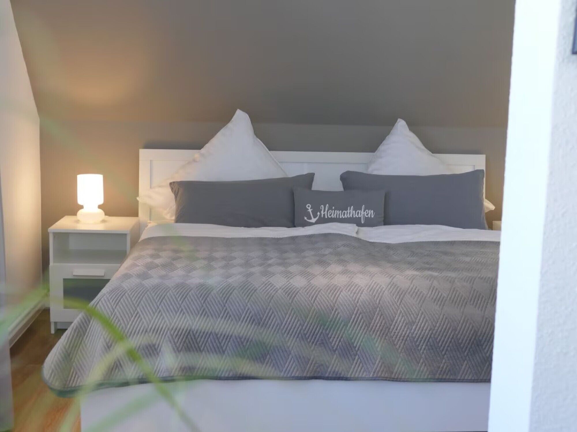 Ein ordentlich bezogenes Bett mit grau-weißer Karodecke, zwei Kissen und einer Nachttischlampe.
