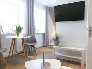 A modern living room with a flat-screen TV, a round table with chairs, a bench, and a potted plant.