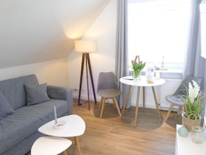 A modern living room with a grey sofa, a small white table, a floor lamp, a dining table with chairs, and a view of the outside.