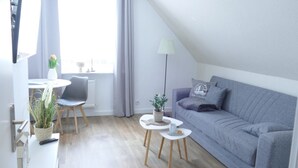 Een moderne, goed verlichte woonkamer met een grijze bank, een klein rond tafeltje, een stoel, een potplant en een lamp.