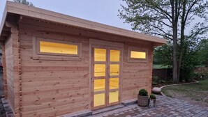 Een houten hutje met een oprit van bakstenen, grote ramen en een kleine tuin.