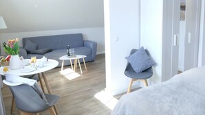 Een moderne, goed verlichte kamer met een grijze bank, een klein rond tafeltje met ontbijtartikelen, een grijze stoel en een witte kast.