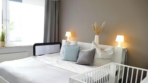 Une chambre à coucher avec un lit blanc, un berceau, deux lampes de chevet, une fenêtre avec des rideaux et une plante en pot.