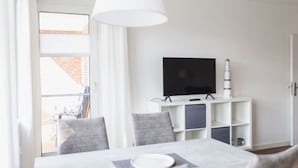 Un salon moderne avec un téléviseur à écran plat posé sur un meuble blanc, une table à manger avec des chaises et une grande fenêtre avec des rideaux.