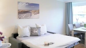 Habitación de hotel con cama, una mesita, una silla, ventana con cortinas y un cuadro en la pared.
