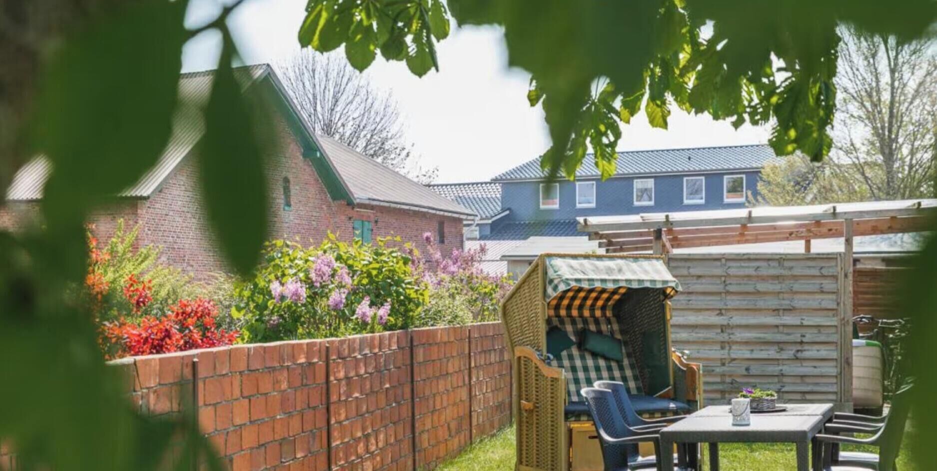 Un jardin à l’arrière de la maison, avec un mur en briques, un petit potager fleuri, une cabane en bois et une table avec des chaises.