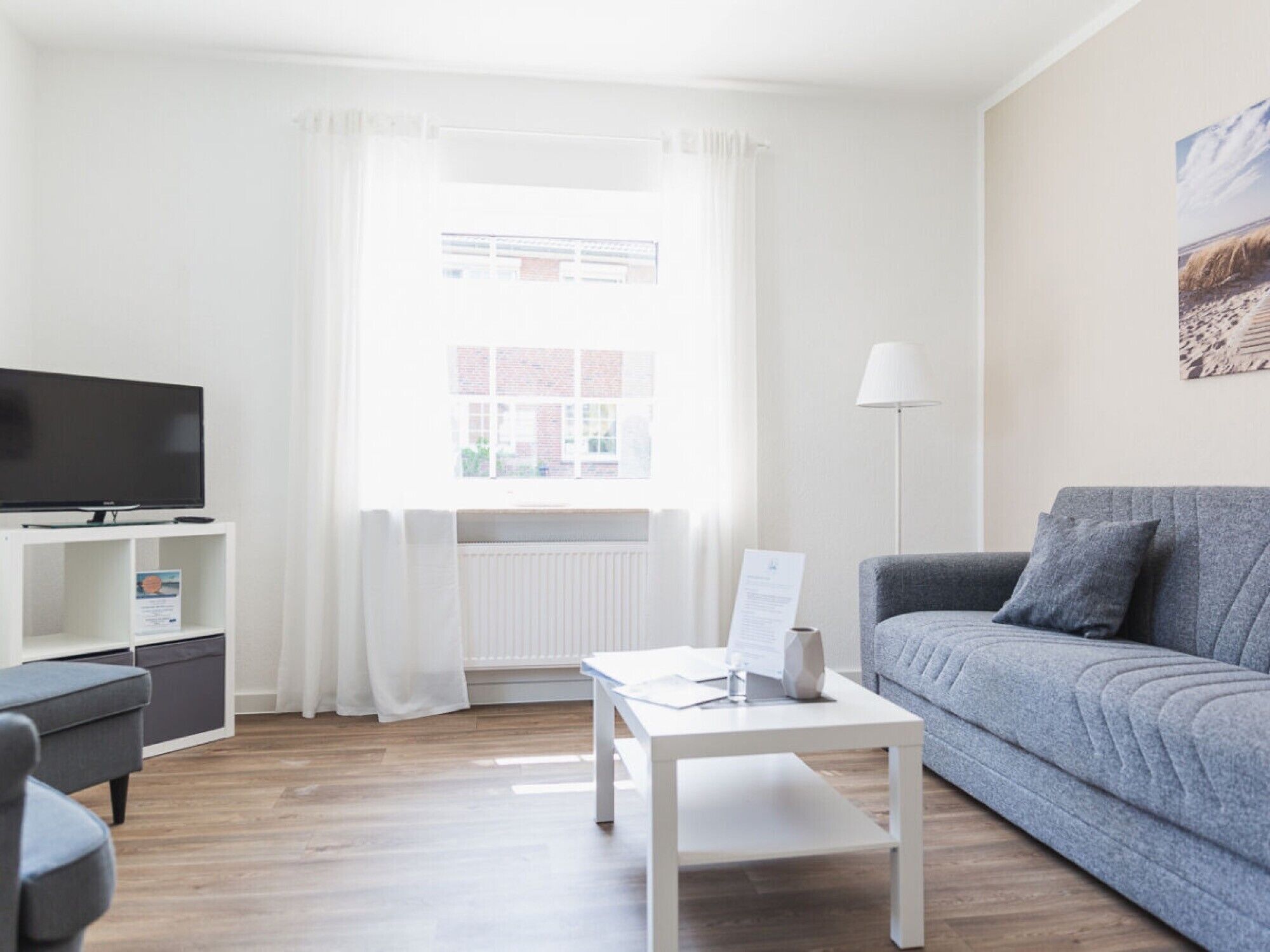 A modern living room with a flat-screen TV, a grey sofa, a white coffee table, and a window with white curtains.