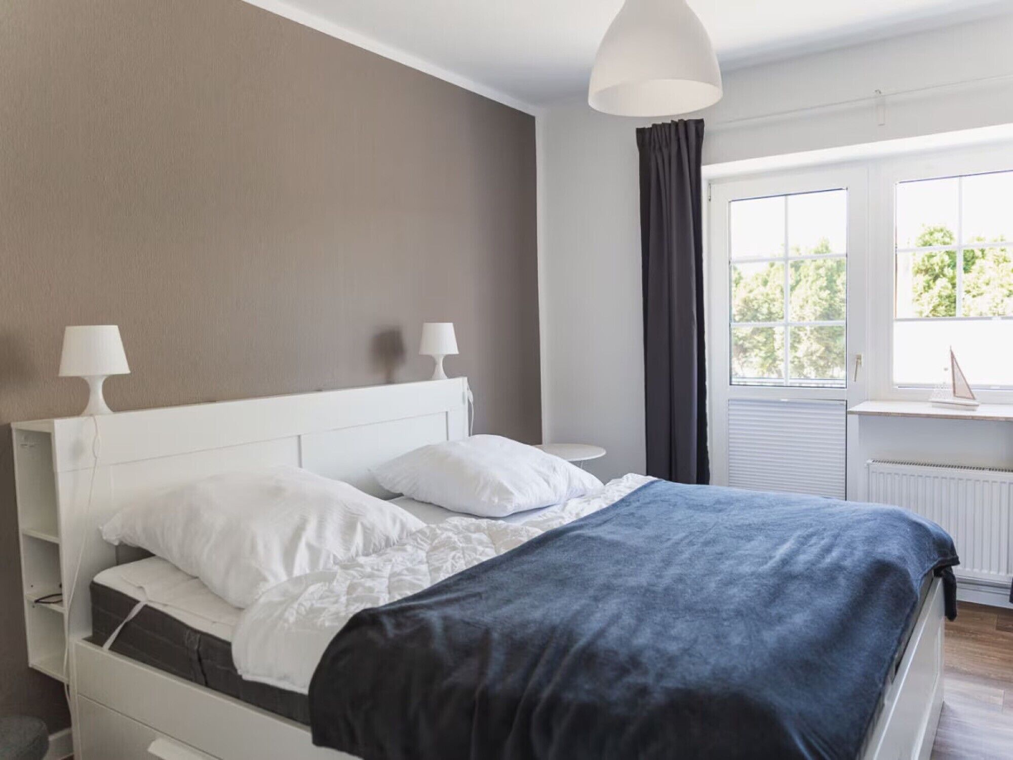Ein Schlafzimmer mit einem Bett, zwei Nachttischlampen, einem Fenster mit Vorhängen und einer Heizung.