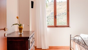 A bedroom with a wooden dresser, a bed with metal headboard, a window with a view of a hilly landscape, and a white curtain. - Room 'Monte Suso' with sea view, shared terrace and air conditioning (Montallegro)