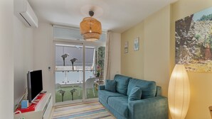 A modern living room with a sofa, a flat-screen TV, a patterned rug, a ceiling fan, and a large window with a view of a balcony.