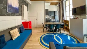 A modern living room with a sofa, a dining table with chairs, a kitchen in the background, and a TV mounted on the wall. - UrbanFlat 44 - Pretty 2 Bedroom Flat in Paris near metro station (París)