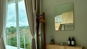 Una habitación con un ventanal grande, un perchero de madera, un espejo y una estantería de madera con artículos de aseo.