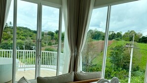 Habitación de hotel con una cama grande, un balcón con baranda blanca y vistas a zonas verdes y colinas.
