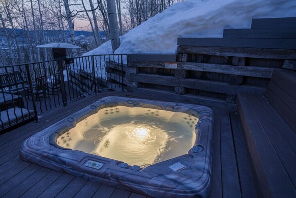 A hot tub on a wooden deck with a metal railing, surrounded by snow and trees.