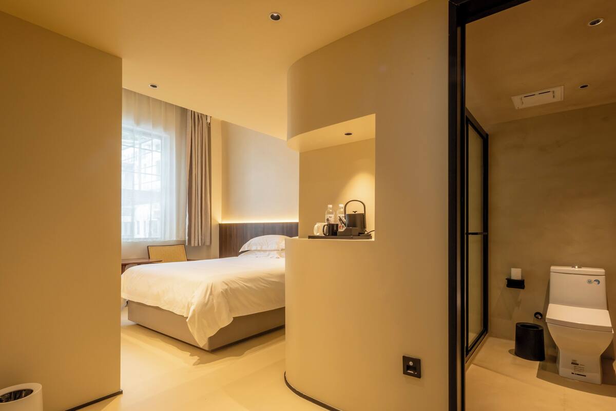 Una habitación de hotel con cama, mesita de noche con lámpara, ventana con cortinas y baño con inodoro y lavamanos.
