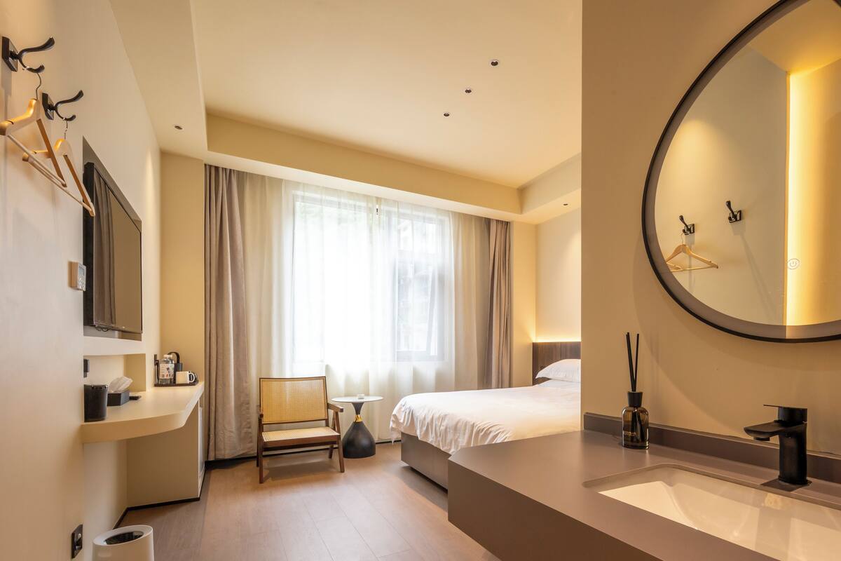 Una habitación de hotel moderna con una cama grande, un espejo redondo, una mesita de noche, una pequeña mesa redonda y una silla.