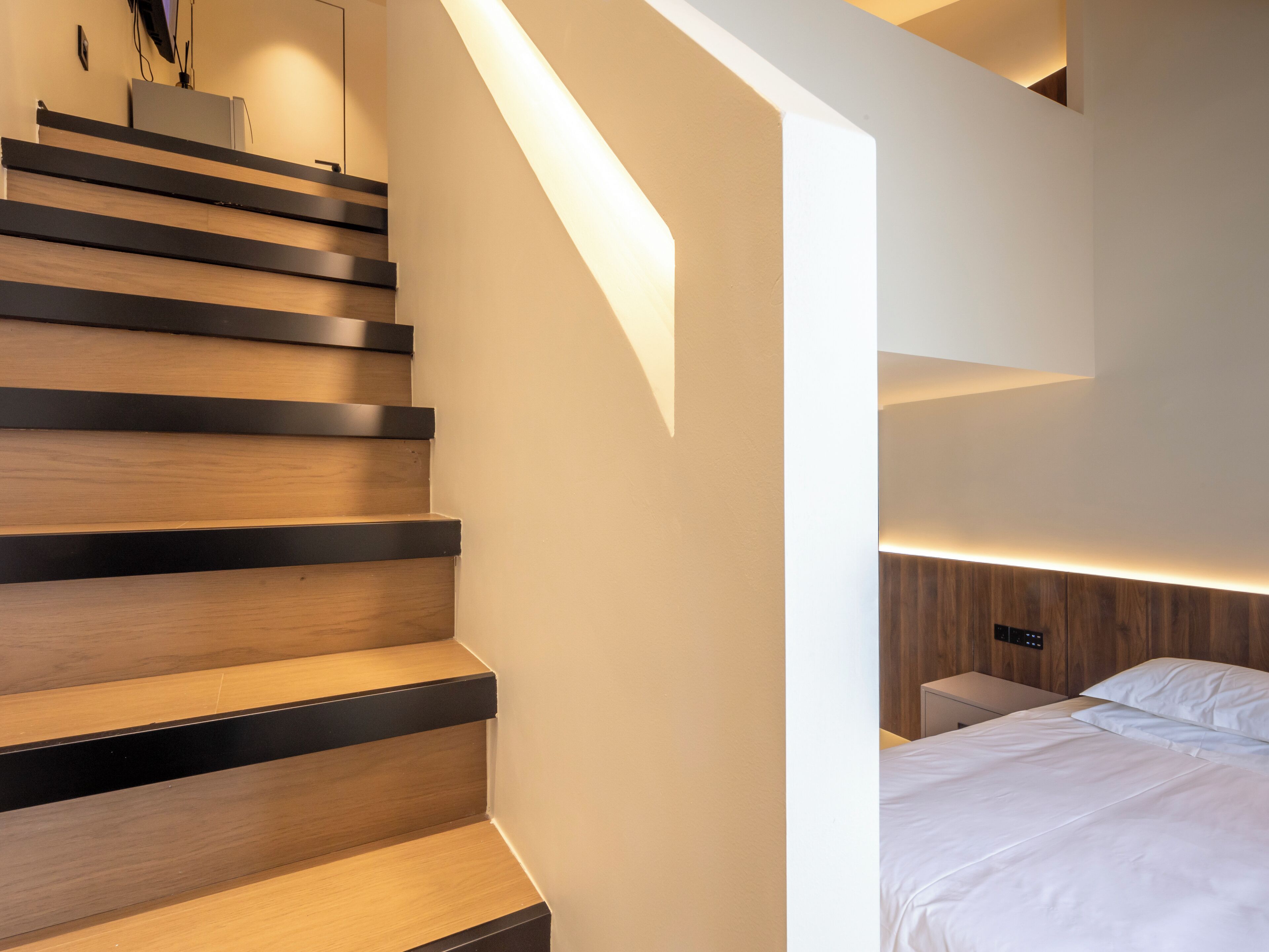 A modern staircase with wooden steps and black edges, leading to a bedroom with a bed and bedside table.