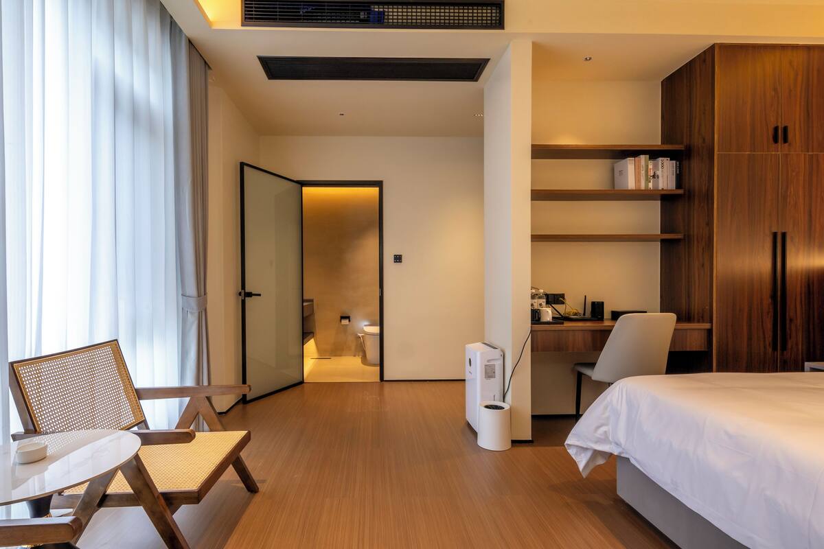 Una habitación de hotel moderna con cama, escritorio, silla, estantería y baño visible a través de una puerta abierta.