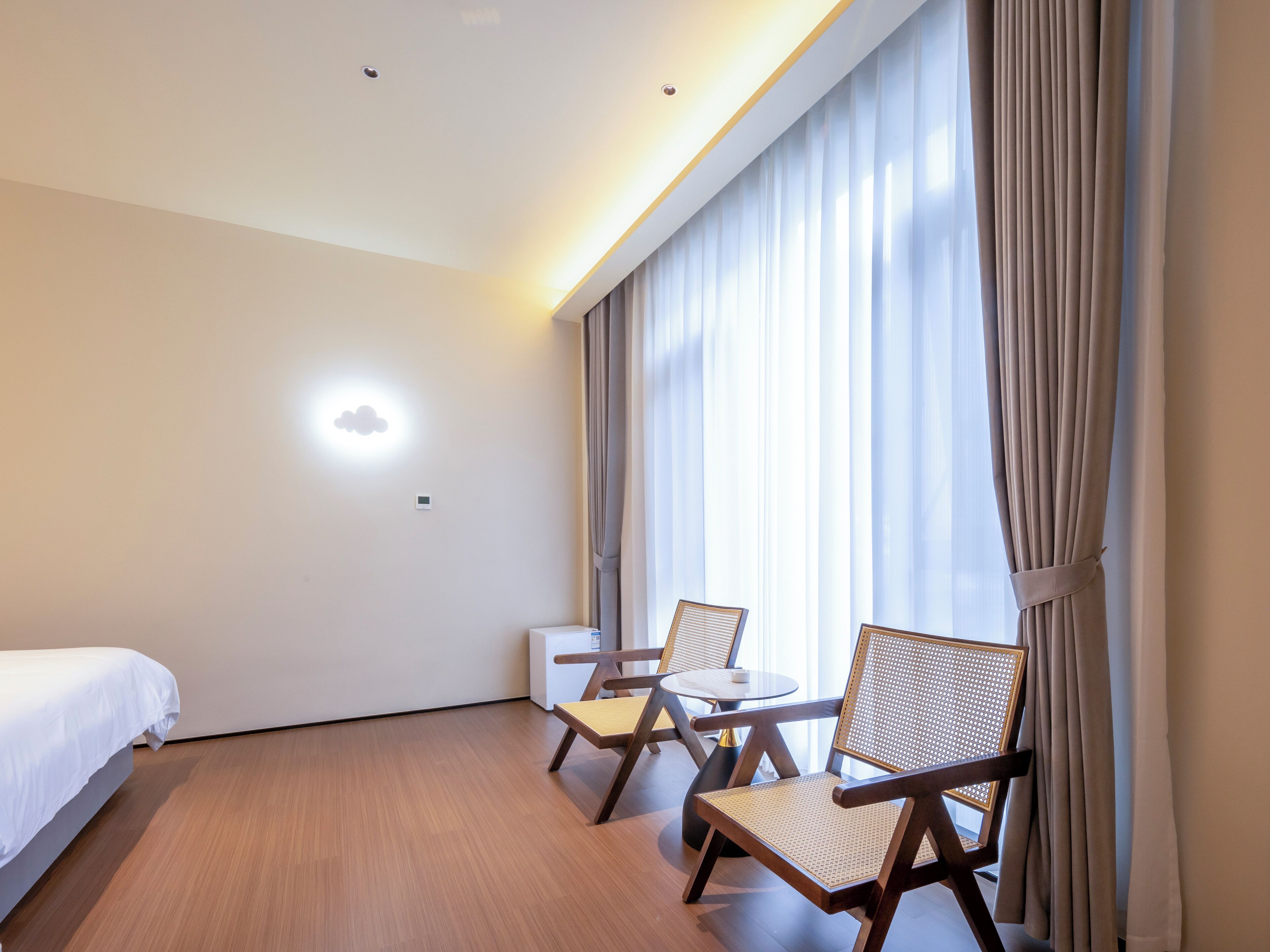 A hotel room with a bed, two chairs, a small table, a wall-mounted air conditioner, and a window with curtains.
