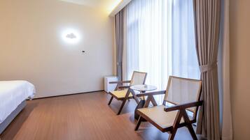 A hotel room with a bed, two chairs, a small table, a wall-mounted air conditioner, and a window with curtains.