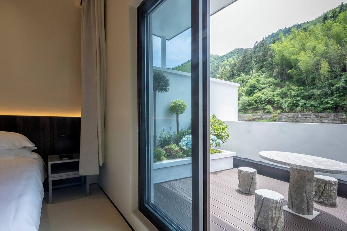 Habitación de hotel moderna con un ventanal grande, una cama, una mesita y vistas a una ladera verde y exuberante.
