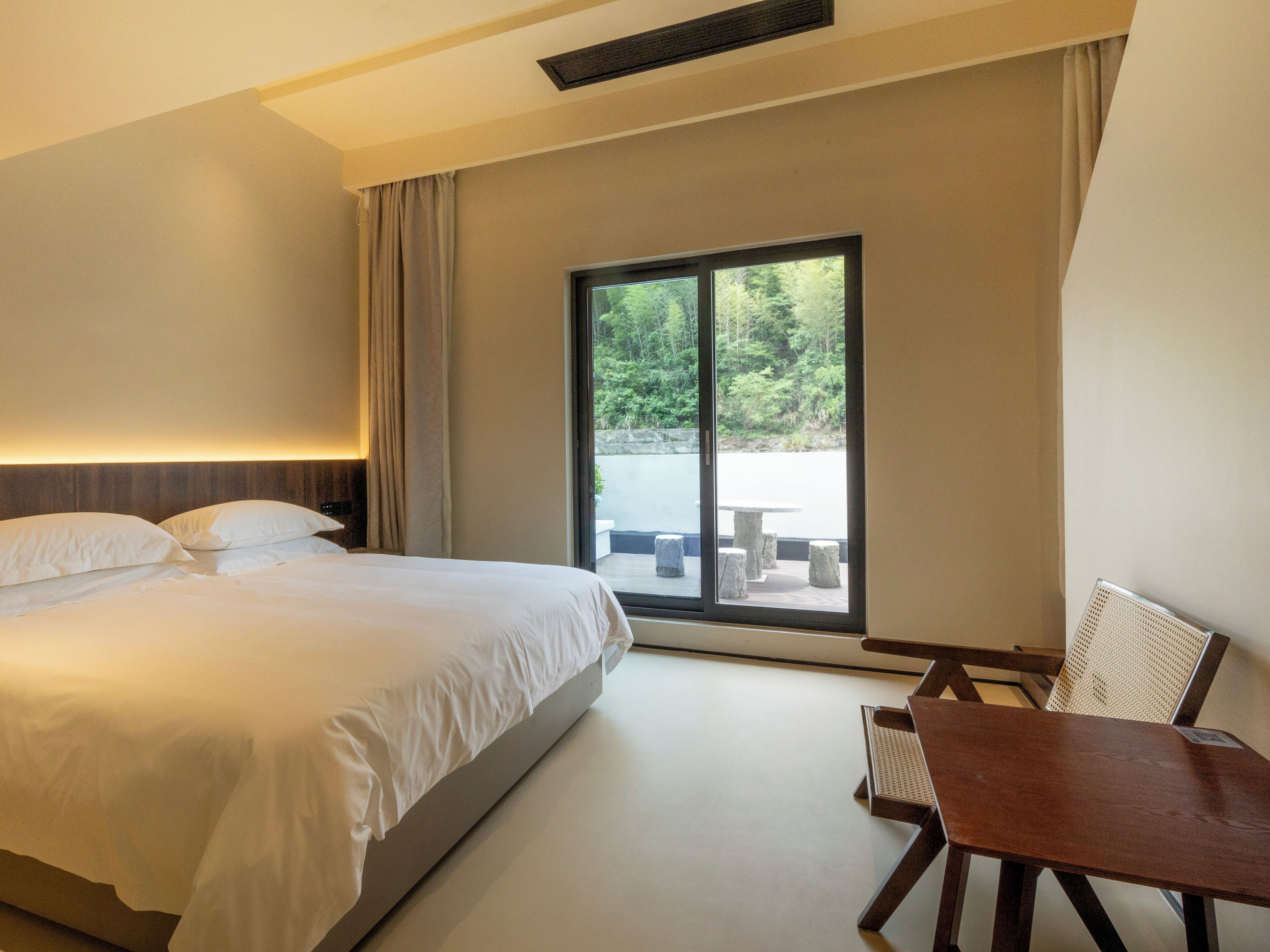 A hotel room with a large bed, a wooden table, and a view of a garden through a sliding glass door.
