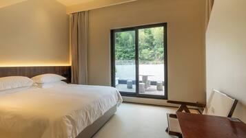 A hotel room with a large bed, a wooden table, and a view of a garden through a sliding glass door.