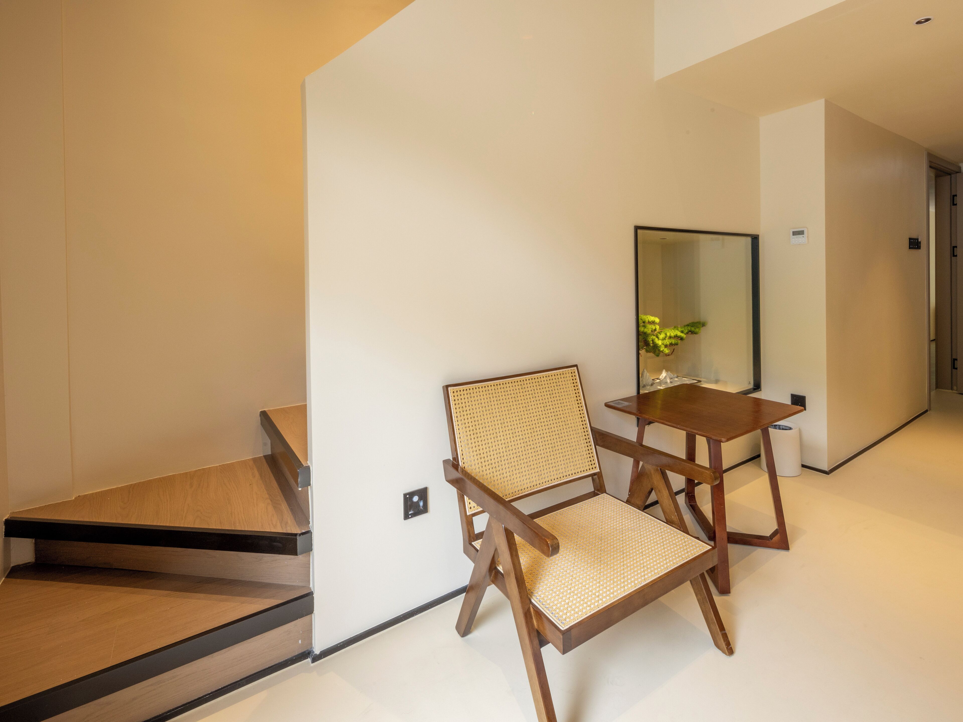 A modern hotel room with a wooden floor, a wooden chair with a woven seat, a small wooden table, and a mirror on the wall.