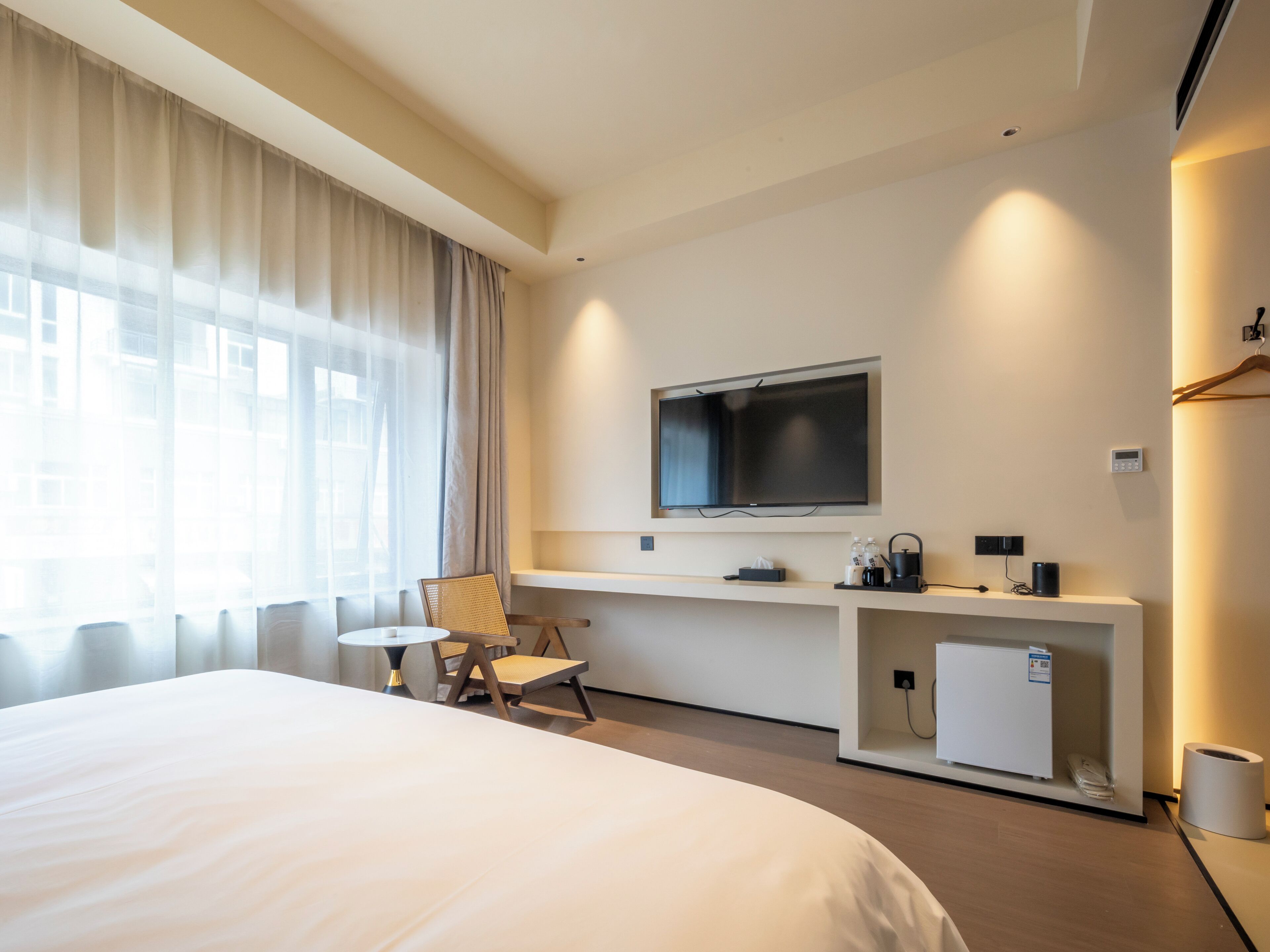 A hotel room with a large bed, a desk with a TV, a chair, a small fridge, and a bedside table with a lamp.