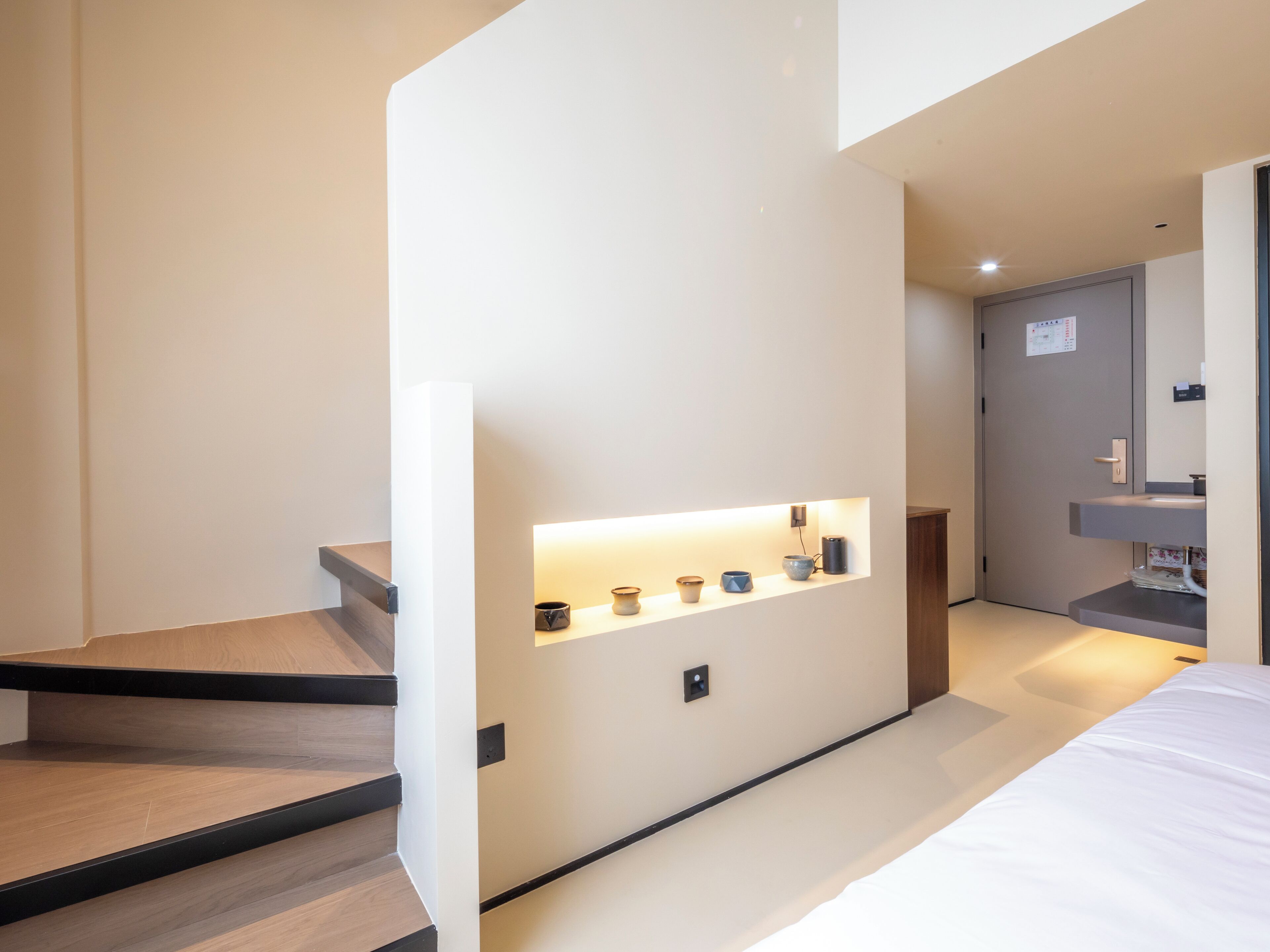 Une chambre d’hôtel moderne avec un escalier en bois, une table de chevet avec une lampe et une petite étagère ornée d’objets décoratifs.