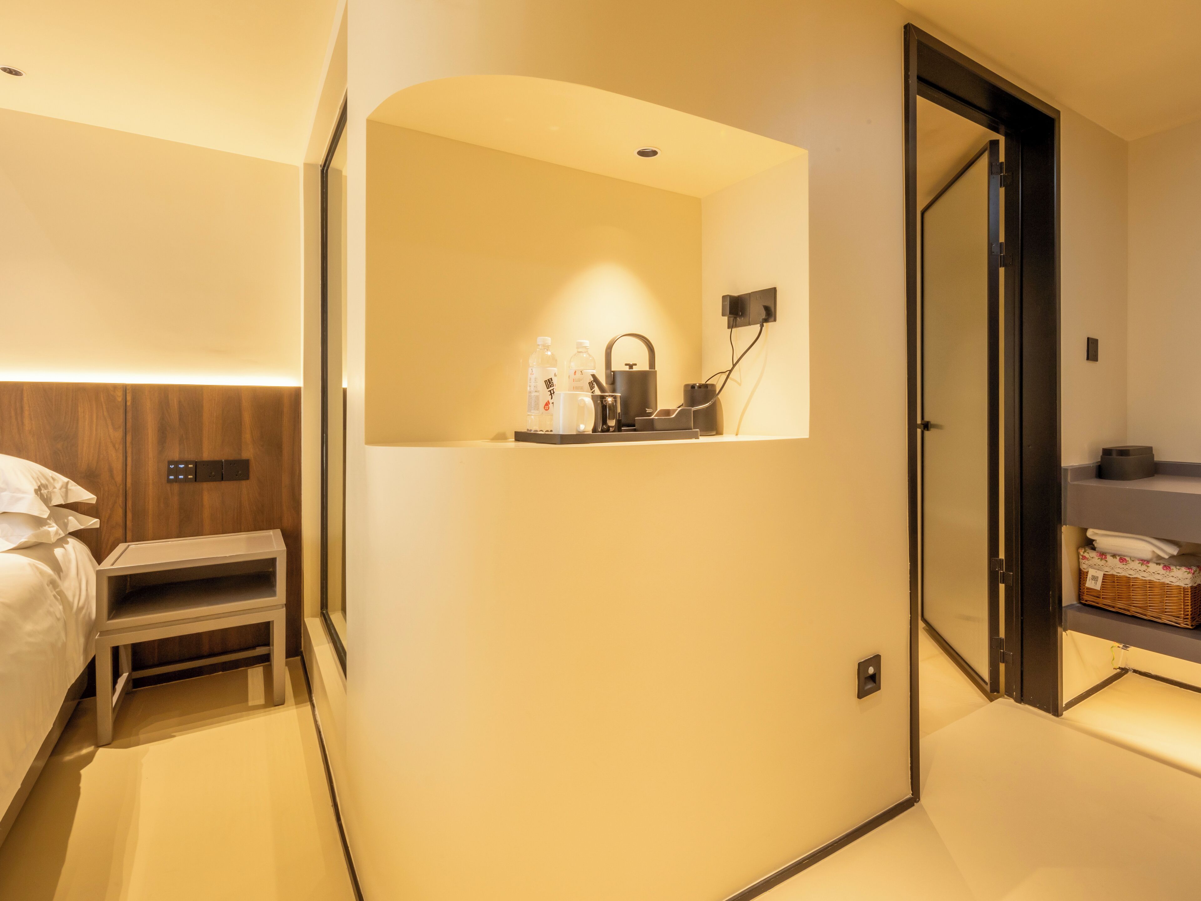 A modern hotel room with a bed, bedside table, and a small desk with a chair. There is a mirror above the desk, and a door leading to a bathroom.