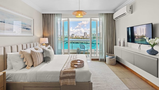 A modern hotel room with a large bed, a television, a balcony with a city view, and an air conditioning unit.