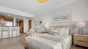 A modern hotel room with a large bed, bedside tables, a desk area with chairs, and a kitchen in the background.