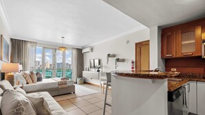 A modern hotel room with a living area, kitchenette, and large windows offering a city view.