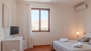 A hotel room with a bed, a desk with a computer, a window with curtains, and an air conditioner.