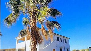 A white building with a balcony, a palm tree in front, and a clear blue sky.